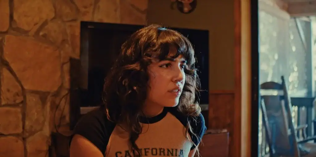 A young woman with curly hair wearing a California t-shirt looks off-camera with a concerned expression.