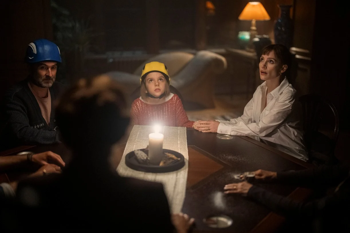 A man, a young girl in a yellow helmet, and a woman sit at a candlelit table with others, looking upward.
