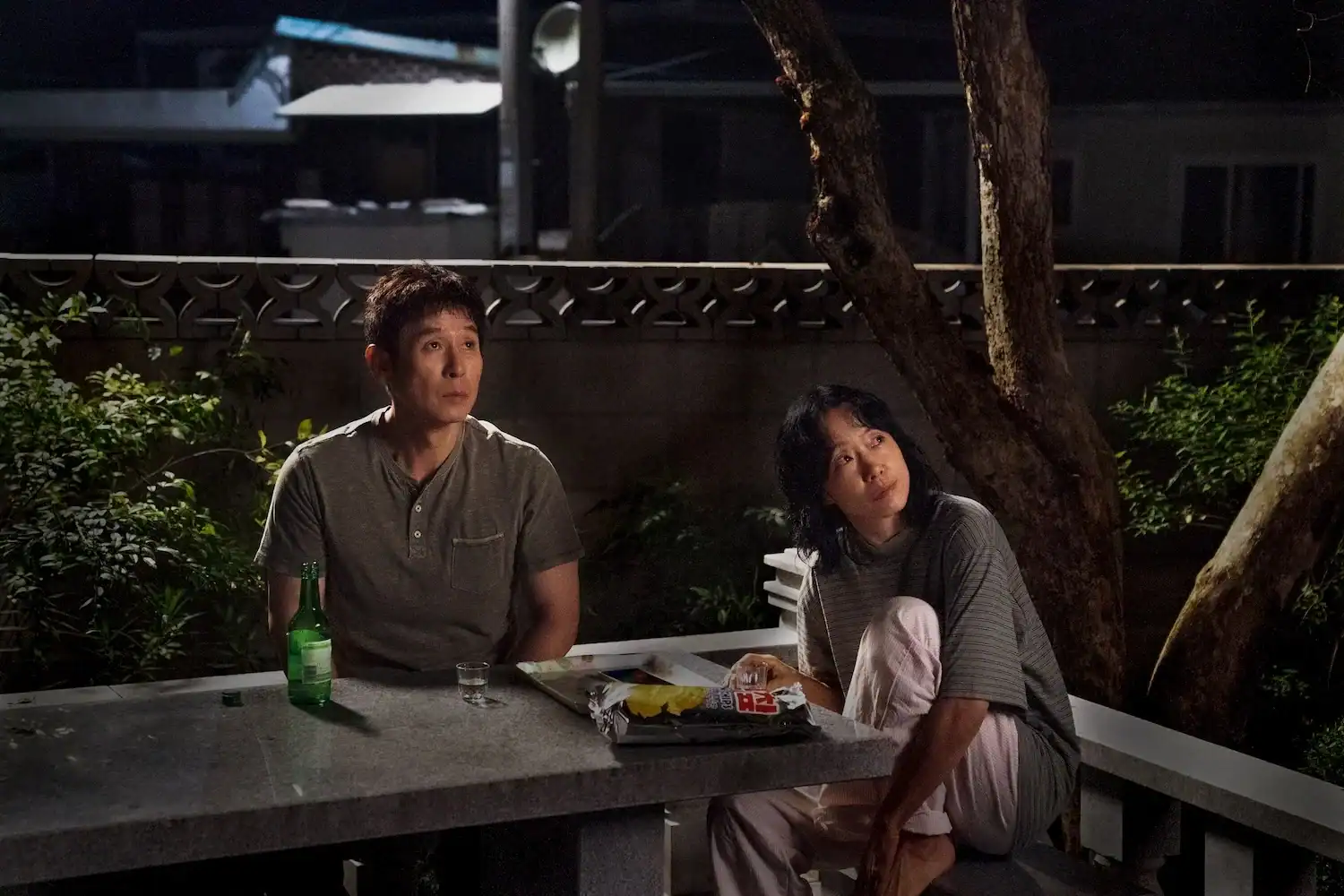 A man and a woman sitting at an outdoor stone table at night with a bottle of soju and snacks.