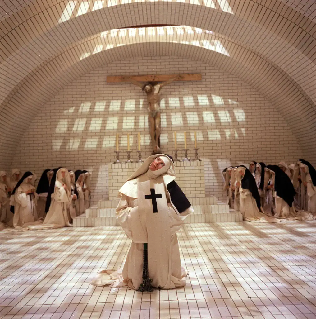 A nun kneels in prayer in the center of a tiled chapel, surrounded by other nuns, with a large crucifix mounted on the wall behind the altar.