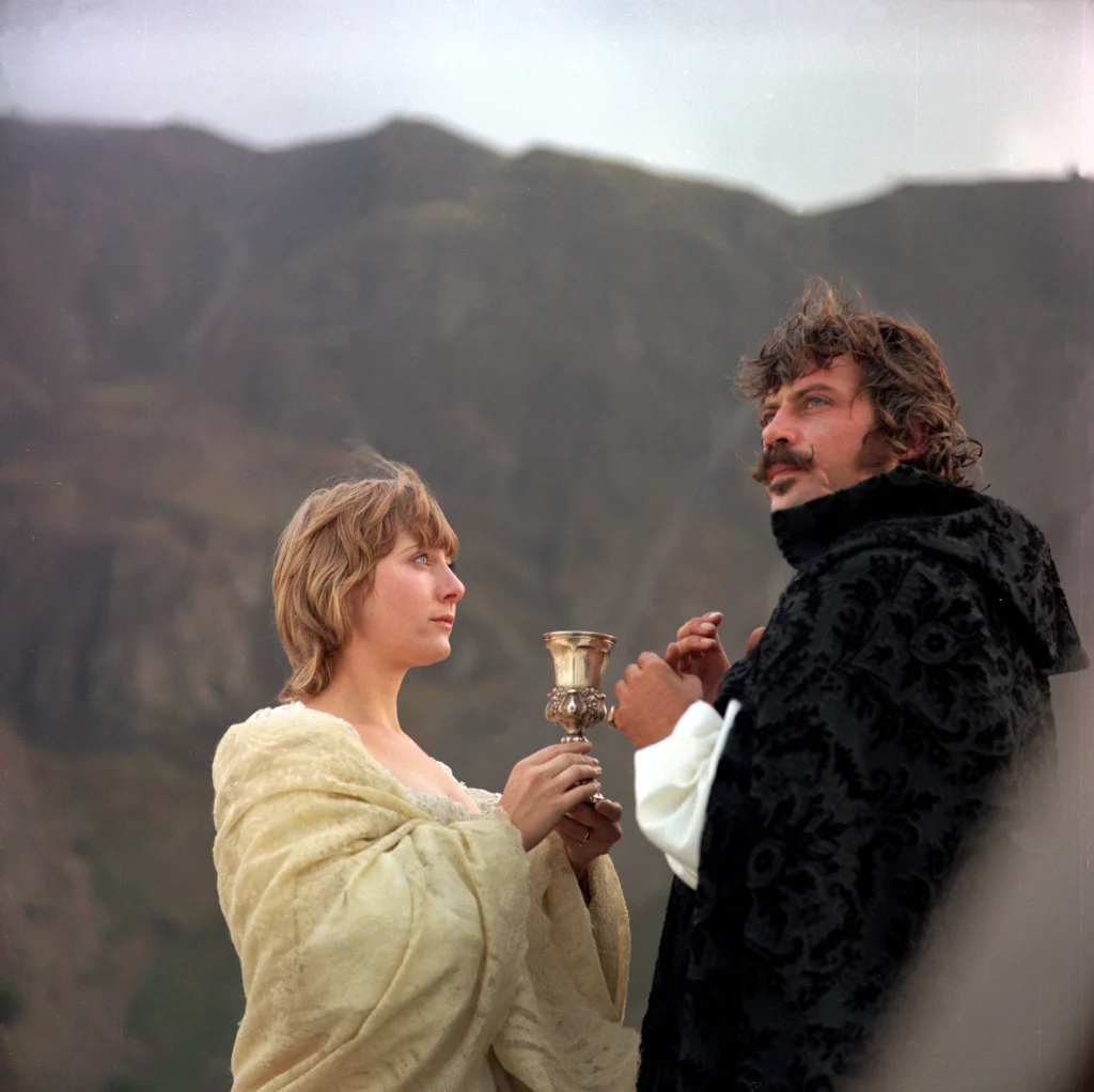 A woman in a cream-colored shawl holds a chalice out toward a man in a black cloak against a backdrop of mountains.