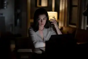 A woman sits at a desk working on a laptop at night, holding a glass of red wine.